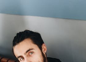 Close-up portrait of a bearded man with green eyes posing indoors against a blue wall.