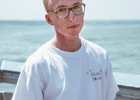 Young man in glasses wearing a white shirt, standing by the sea on a sunny day.