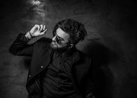 Black and white portrait of a fashionable man in sunglasses and suit posing confidently indoors.